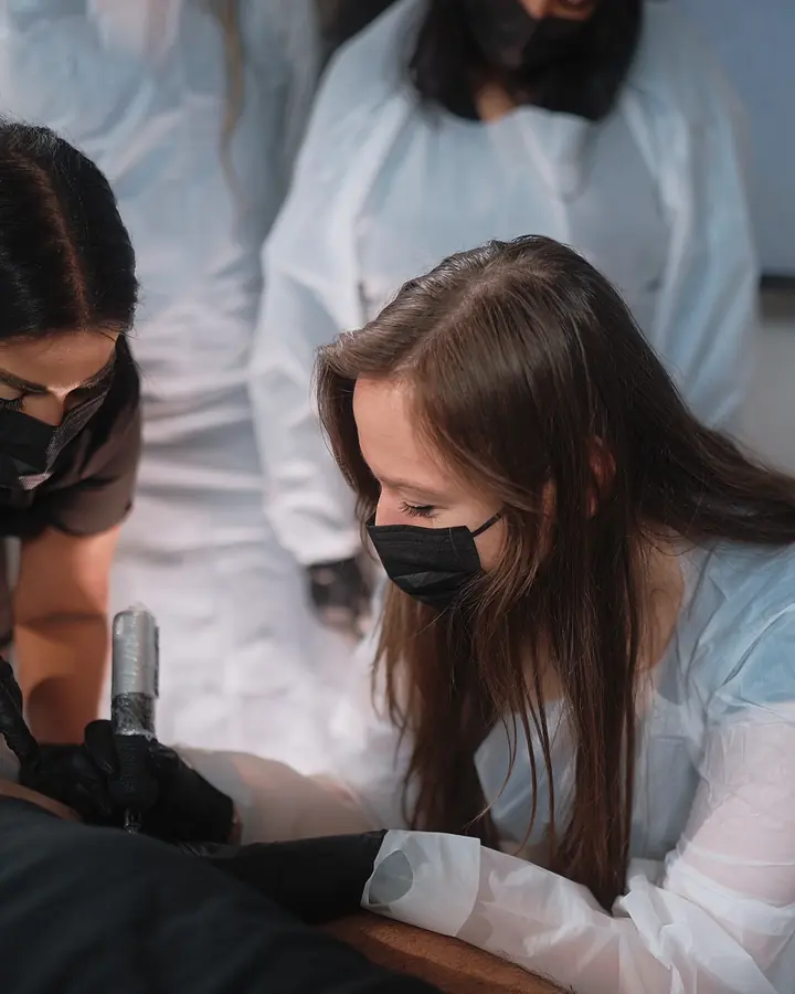 An Academy student, masked and gloved, working on a live model’s leg while two educators watch closely